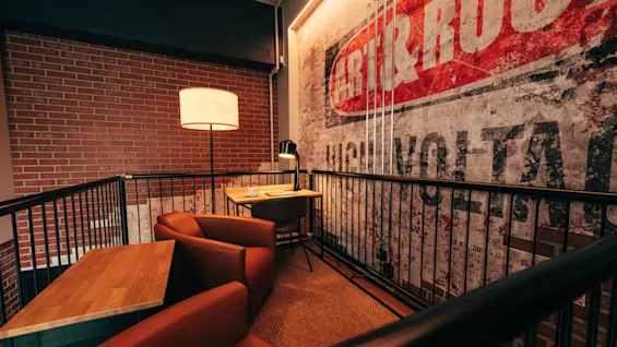 A cozy lounge area with comfortable seating, a wooden table, and lamps, set against a brick wall and a distressed mural with text ART & ROC HIGH VOLTA.