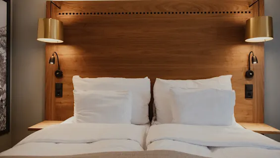 En hyggelig hotelseng med sengegavl i træ, hvidt sengetøj og varm belysning fra to sengelamper, der indbyder til afslapning.
