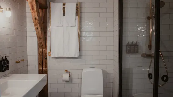 Et moderne badeværelse med hvide fliser, håndvask, toilet og bruser. Hvide håndklæder hænger på en gylden håndklædetørrer, og en rustik træbjælke tilføjer karakter.