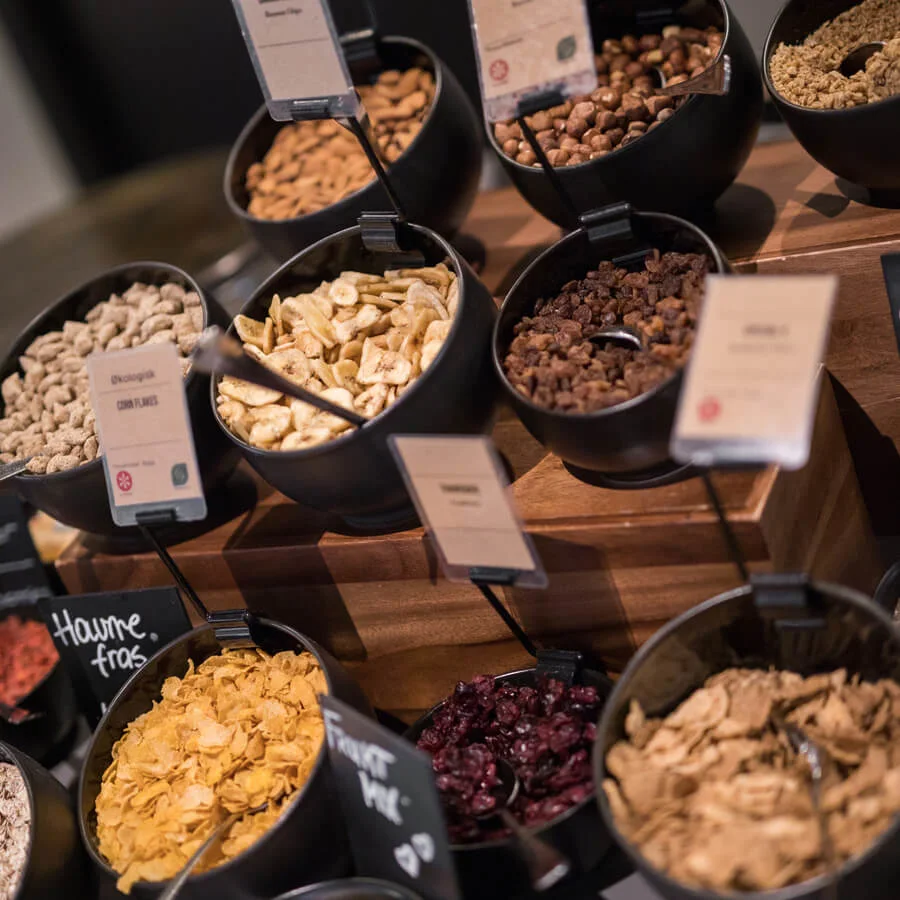 Skålformade behållare innehåller olika nötter och torkad frukt märkta med kort. Placerade på trähyllor bildar de en självbetjäningsstation. Etiketter inkluderar "Corn Flakes" och "Farmors Müsli".