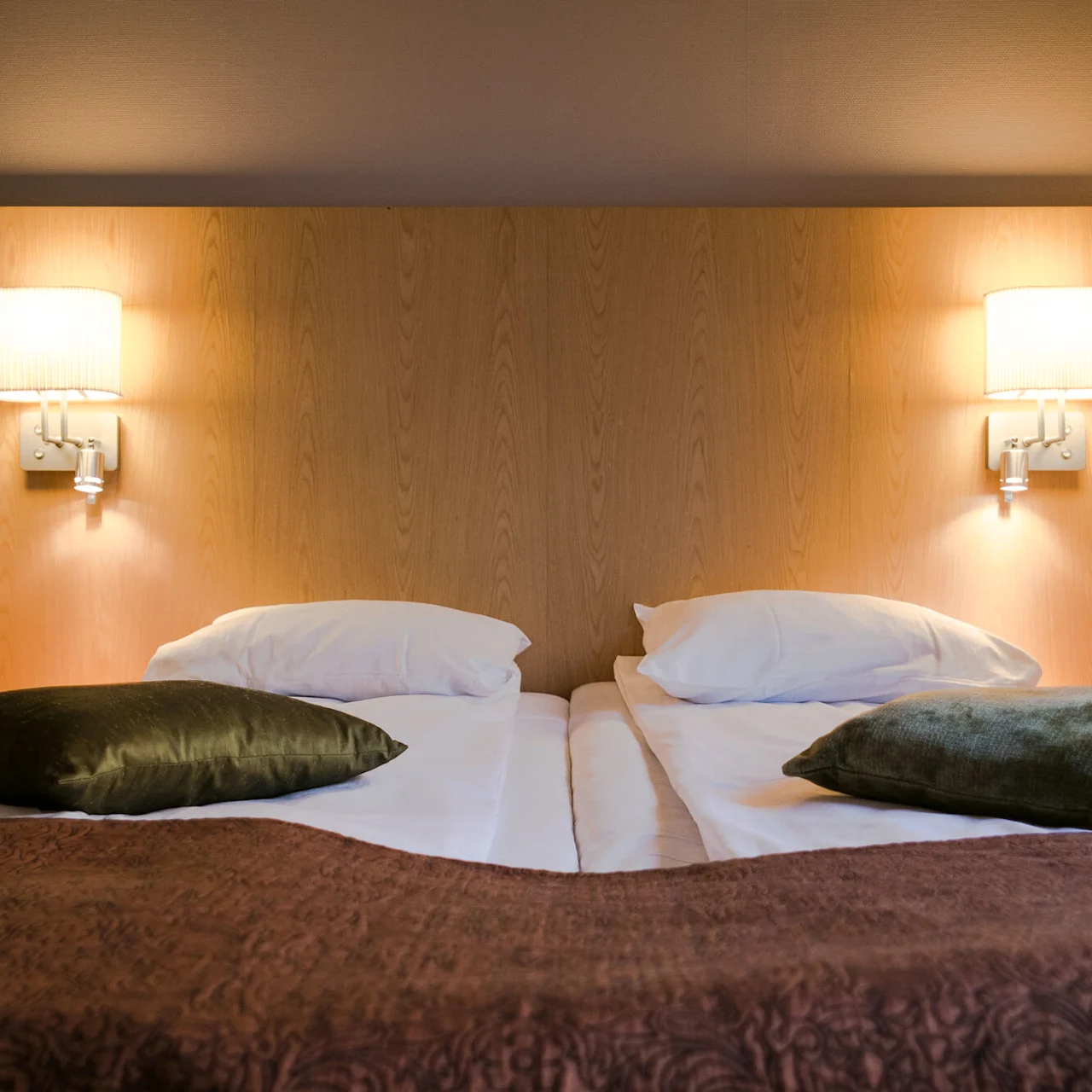A neatly made double bed with two pillows and green cushions is flanked by wall-mounted lamps on a wooden headboard, set in a softly lit bedroom.