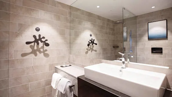 Sink with a faucet reflecting in a bathroom mirror, surrounded by beige tiled walls. Towels hang on a rack nearby, and abstract wall art is visible.