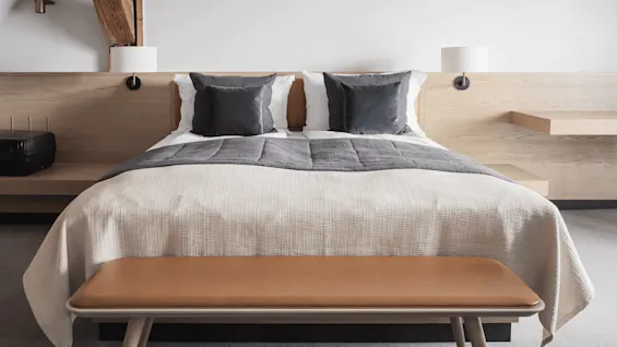 Bed with gray and white linens, two pillows, and a wooden headboard; flanked by wall-mounted lamps. A suitcase is on the left. Modern bedroom with light wood features.