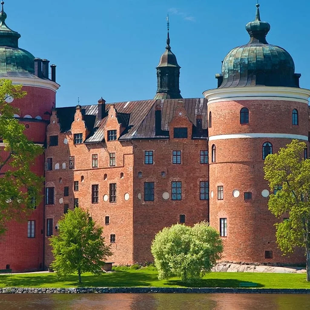 Et storslået murstensslot med grønne kuppelformede tårne står ved et roligt vand, omgivet af frodige grønne træer under en klarblå himmel.
