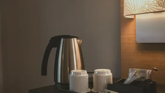Electric kettle sits unused on a table beside two white cups and a tray of tea supplies, under a decorative lamp in a cozy indoor setting.