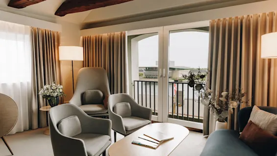 Elegant hotellsvit med en mysig sittgrupp, synliga takbjälkar och en fransk balkong med utsikt.