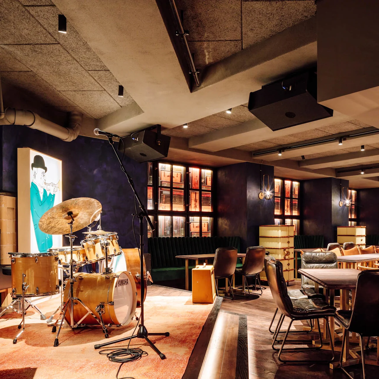 A drum set with a microphone stands on a small stage in a cozy, dimly lit restaurant with modern decor and plush seating.