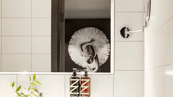 A bathroom sink with two soap dispensers labeled "Z" sits below a mirror reflecting a ballerina in white. A small plant and white towels rest on the wooden counter.