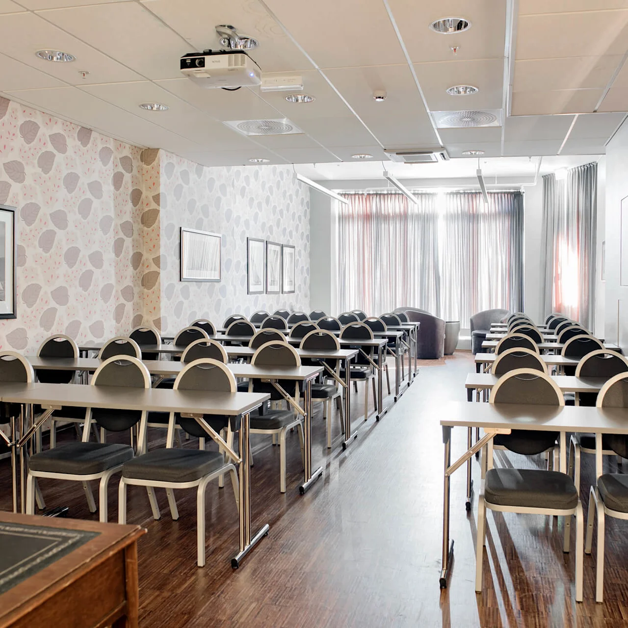 Rader av bord og stoler i et konferanserom med mønstret tapet, bilder på veggen, og gardiner for vinduene.