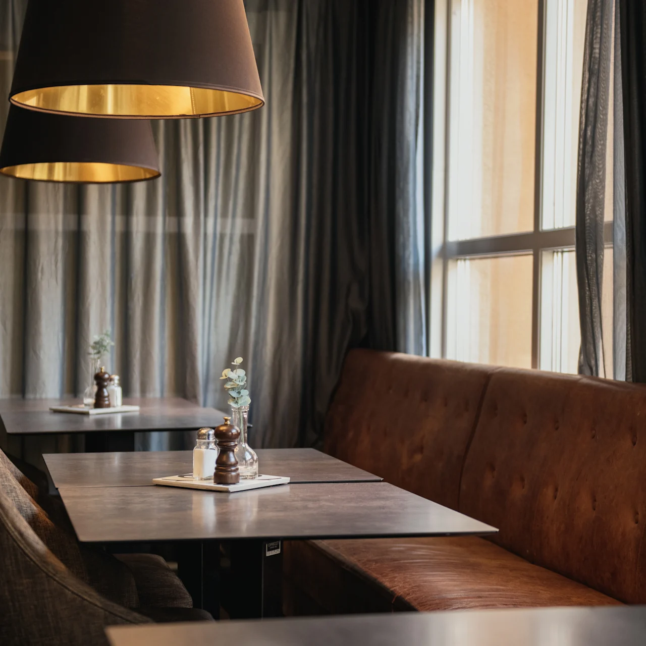 Cozy cafe setting features brown leather seating and wooden tables adorned with salt, pepper, and flower vases. Large, elegant lampshades hang above, with sheer curtains and a large window providing natural light.