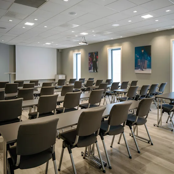 Et moderne konferanserom med rader av bord og stoler vendt mot en stor projeksjonsskjerm, klart for en presentasjon eller et møte.