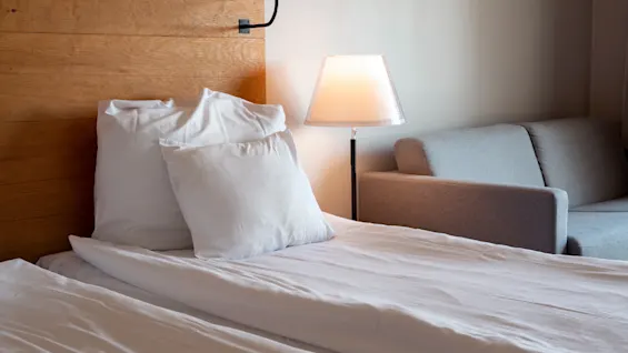 Et hyggeligt hotelværelse med en seng med hvidt sengetøj, en sengegavl i træ, en varm lampe og en komfortabel sofa.