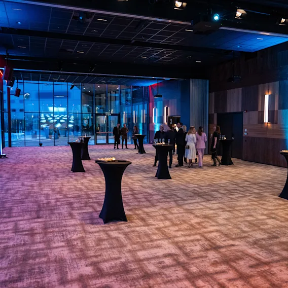 En romslig teppebelagt sal har høye cocktailbord og noen mennesker som står og snakker ved inngangen. Rommet er dempet opplyst med blått og rødt lys, noe som skaper en elegant atmosfære.