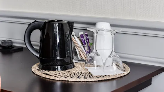 Electric kettle rests on a woven mat, accompanied by inverted wine glasses and assorted tea sachets, on a dark wooden table against a light wall.