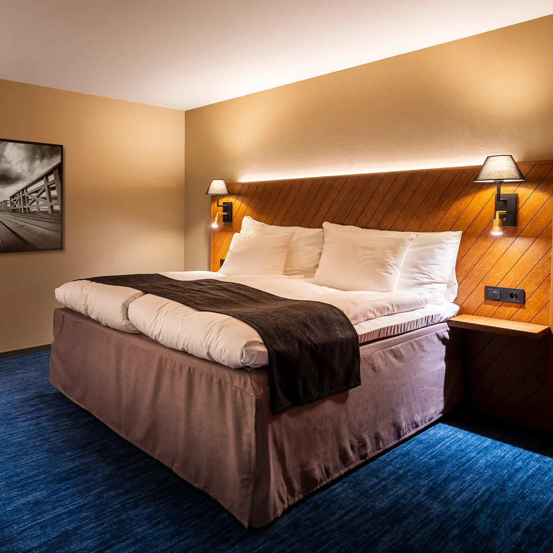 Ett välbelyst hotellrum med en prydligt bäddad säng, konstverk och möbler i en samtida stil.