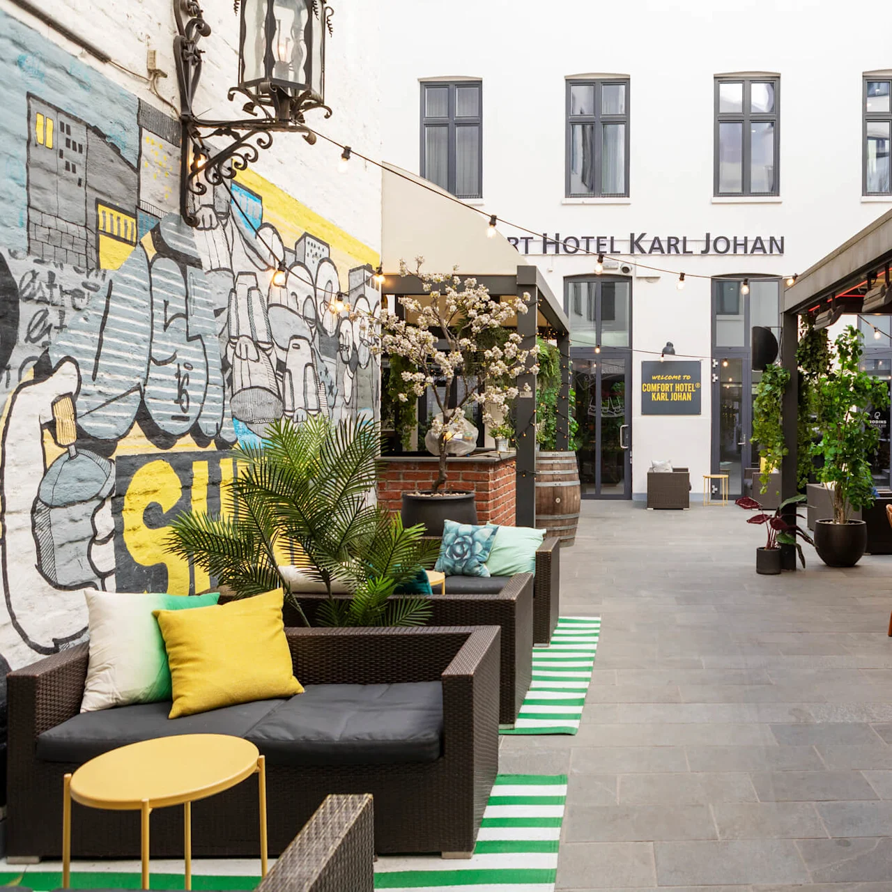 Sittegruppe med rottingmøbler og fargerike puter, ved siden av en veggmaleri. Kontekst inkluderer potteplanter og et skilt: "WELCOME TO COMFORT HOTEL KARL JOHAN."