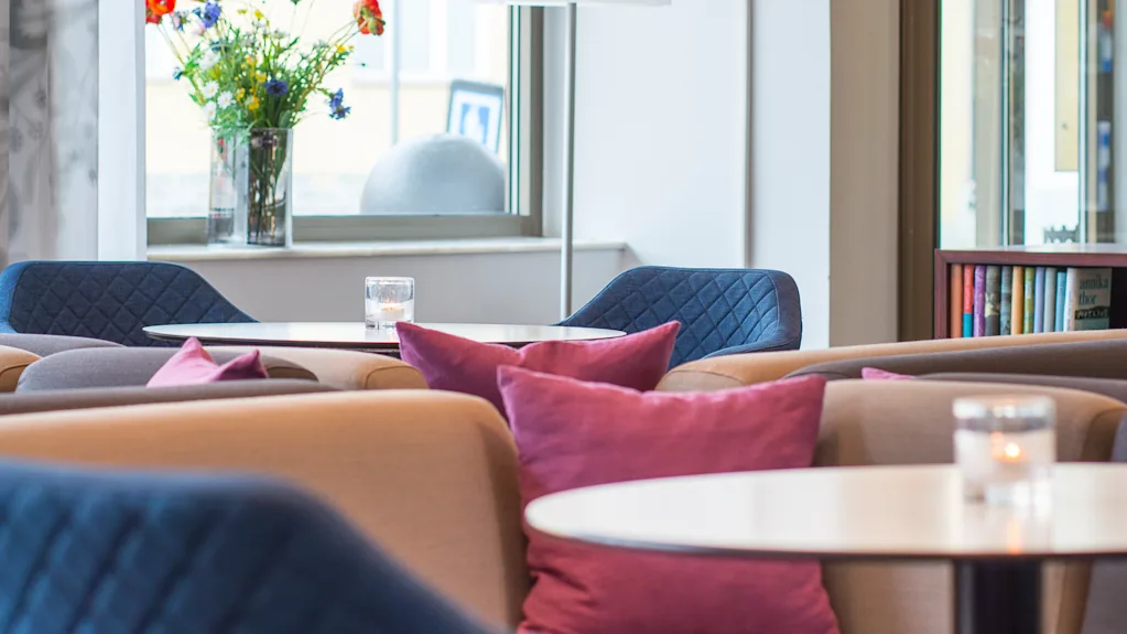 Komfortabel sittegruppe i et moderne oppholdsrom med blå og beige stoler, lilla puter og et bord, omgitt av store vinduer og en vase med fargerike blomster.