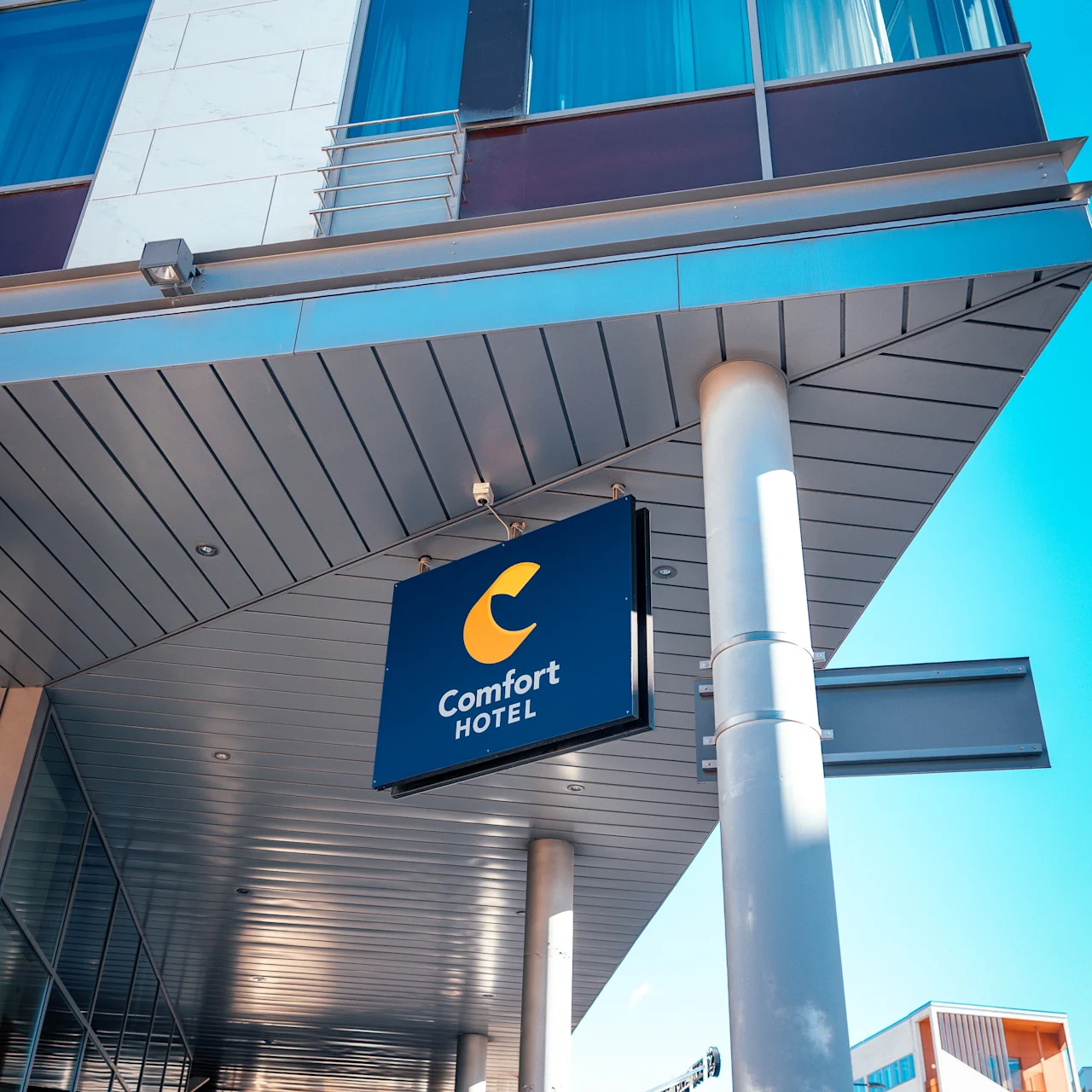 Sign hanging under building canopy, displaying "Comfort HOTEL" in white on blue background, part of a modern glass and metal structure, against a clear blue sky in an urban setting.