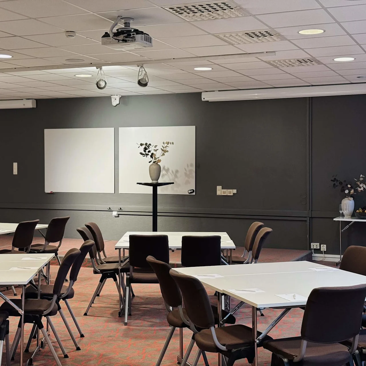 Conference room with arranged tables and chairs, equipped with a projector. Whiteboards hang on a dark wall. Decorative vases with foliage and snacks are on a side table.