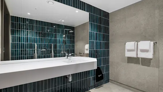 Et moderne bad med hvit vask, stort speil og dusj med mørkeblå fliser. Friske håndklær og en pose merket Hair er synlige.