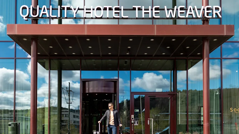 A man with a suitcase exits a glass-fronted hotel under a sign reading "QUALITY HOTEL THE WEAVER" on a sunny day reflected in the windows.