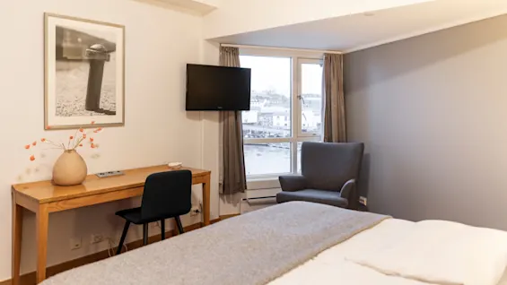 Et koselig hotellrom med en seng, et skrivebord, en TV og en lenestol ved et vindu med utsikt over en by.