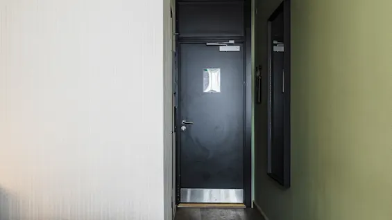 Et koselig hotellrom med minimalistisk dekor, en mørk tredør, myk belysning og en seng med hvit dyne og et oransje pledd som skaper en varm atmosfære. En smal gang fører til utgangen.