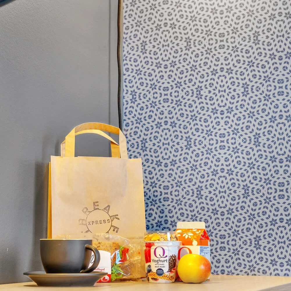Paper bag labeled "XPRESS," surrounded by a coffee cup, pastries, yogurt labeled "Q Yoghurt med nøkkelhull", an apple, and juice carton. Placed on a wooden table, against a patterned wall.