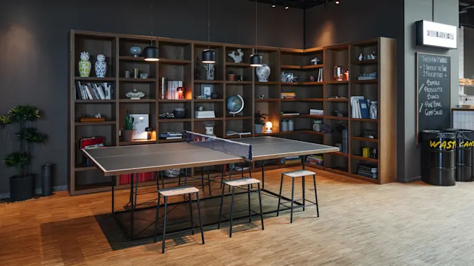 Ping pong table with stools is set in a modern room, surrounded by bookshelves filled with books and decorative items. On the wall: “FOOD PICK-UP”, “THIS IS HOW IT WORKS 1 ORDER AT THE BAR 2 PICKUP FOOD BUFALO, PROSCIUTTO, BIANCO, POKE BOWL, GUMP SALAD".