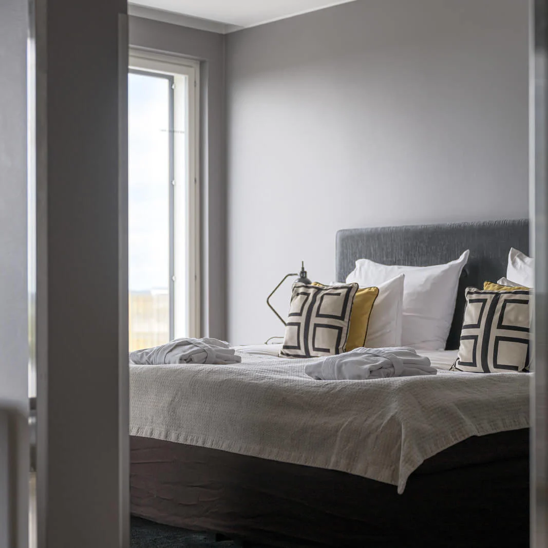 A neatly made bed with geometric-patterned pillows and folded robes is positioned under a large window, providing natural light in a modern, gray-walled bedroom.