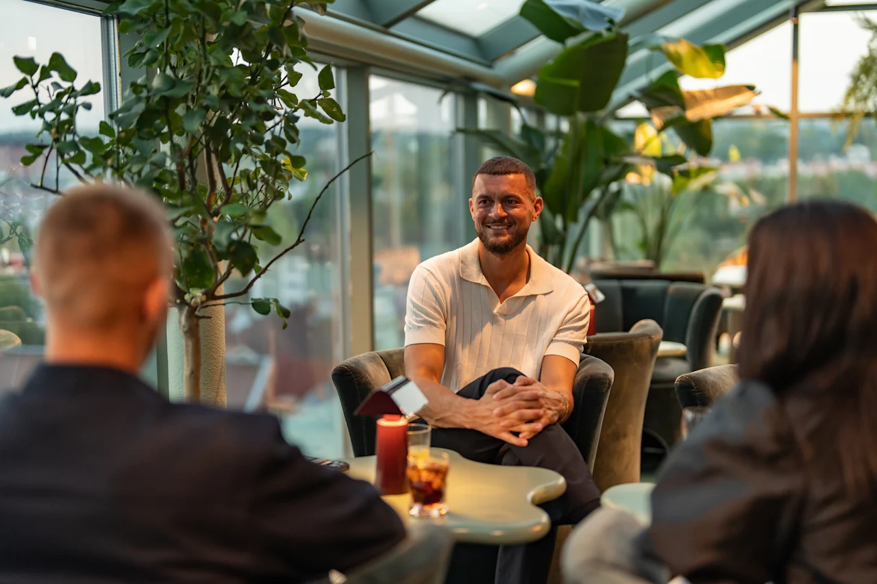 Ihmiset vilkkaassa keskustelussa valoisassa, kasveilla täytetyssä oleskelutilassa, nauttien juomia ja toistensa seurasta.