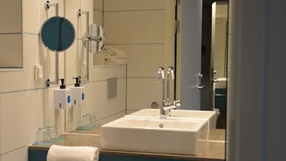 En hvid vask med en kromvandhane er centreret på en blå bordplade. I nærheden er to glaskopper, vægmonterede spejle og håndklædedispensere i et flisebelagt hotelbadeværelse.