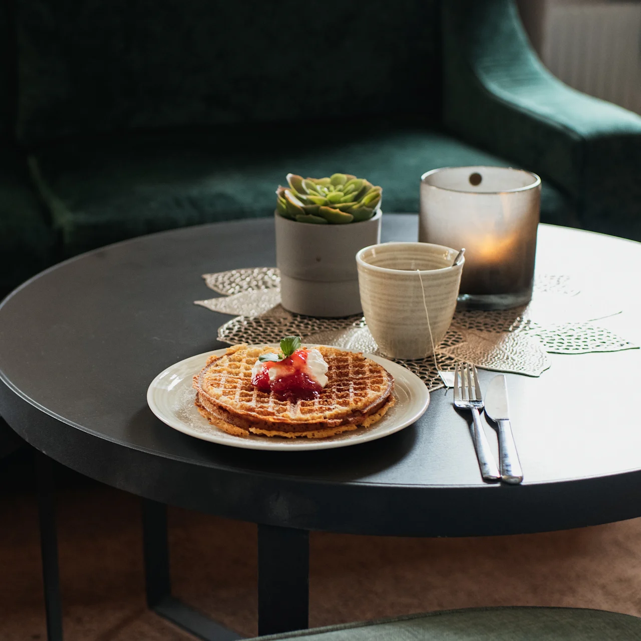 Waffles topped with cream and strawberries rest on a plate beside cutlery and a cup of tea on a round table. A succulent and lit candle decorate the cozy, dimly lit room.