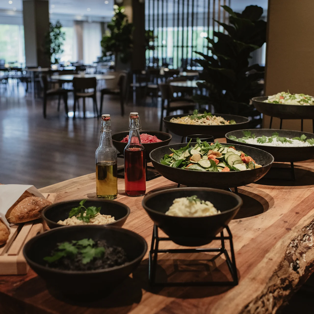 Assorted dishes on a wooden buffet table hold salads, bread, and condiments. This spread is set in a spacious, elegantly furnished dining area, with tables and chairs in the background.