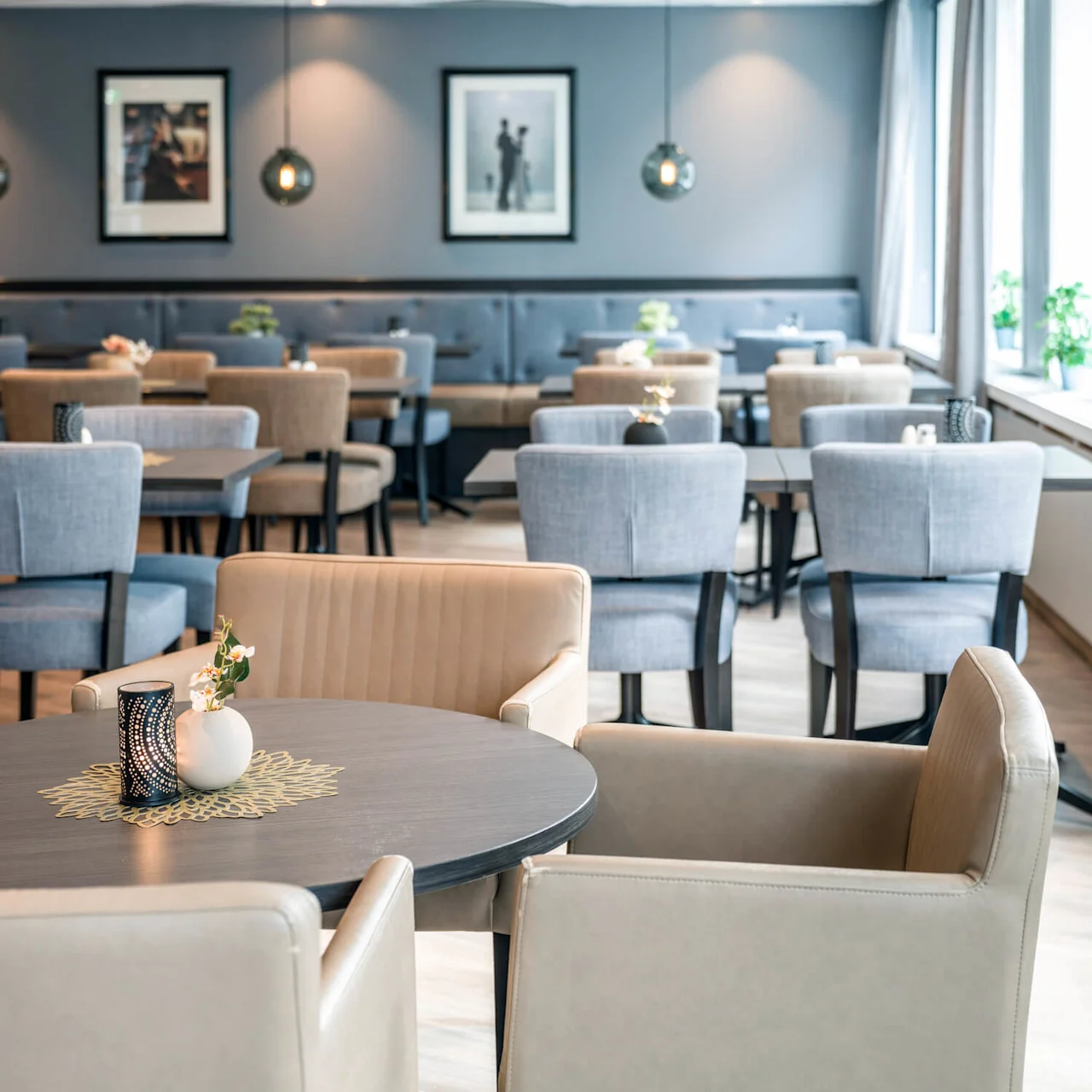 Tables with vases and chairs are arranged in a well-lit, modern dining room with framed photos on gray walls and large windows.