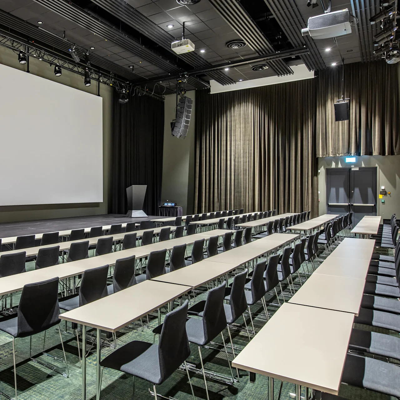Rows of empty black chairs and white tables face a large white projection screen. Curtains and overhead speakers are present in a spacious conference room setting.