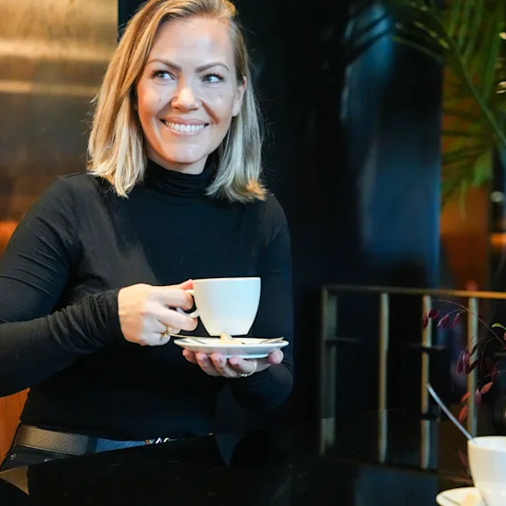Hymyilevä vaaleahiuksinen nainen mustassa poolopaidassa istuu tumman pöydän ääressä kotoisassa loungessa pitelemässä valkoista kahvikuppia.