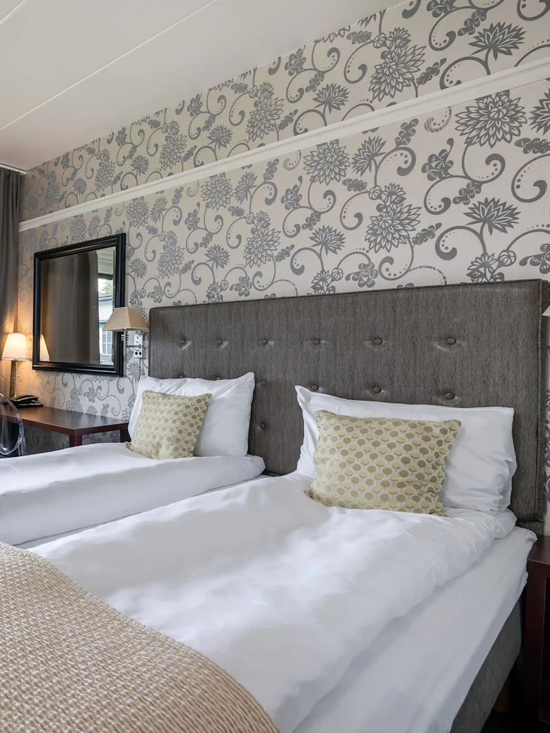 A neatly made twin bed with white linens and patterned cushions sits in a room with floral wallpaper, a window, a brown chair, and a nightstand with a lamp.