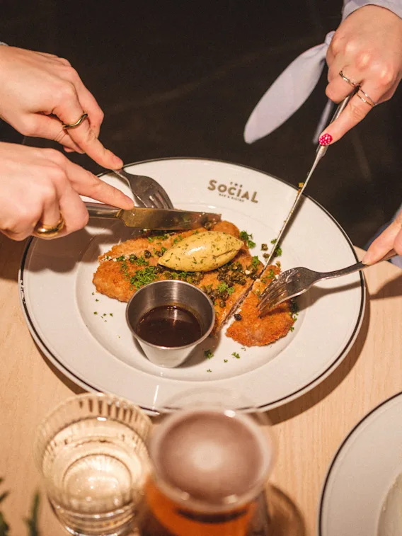 Hands cutting a shared meal on a plate with the word SOCIAL, enjoying a dining experience together.