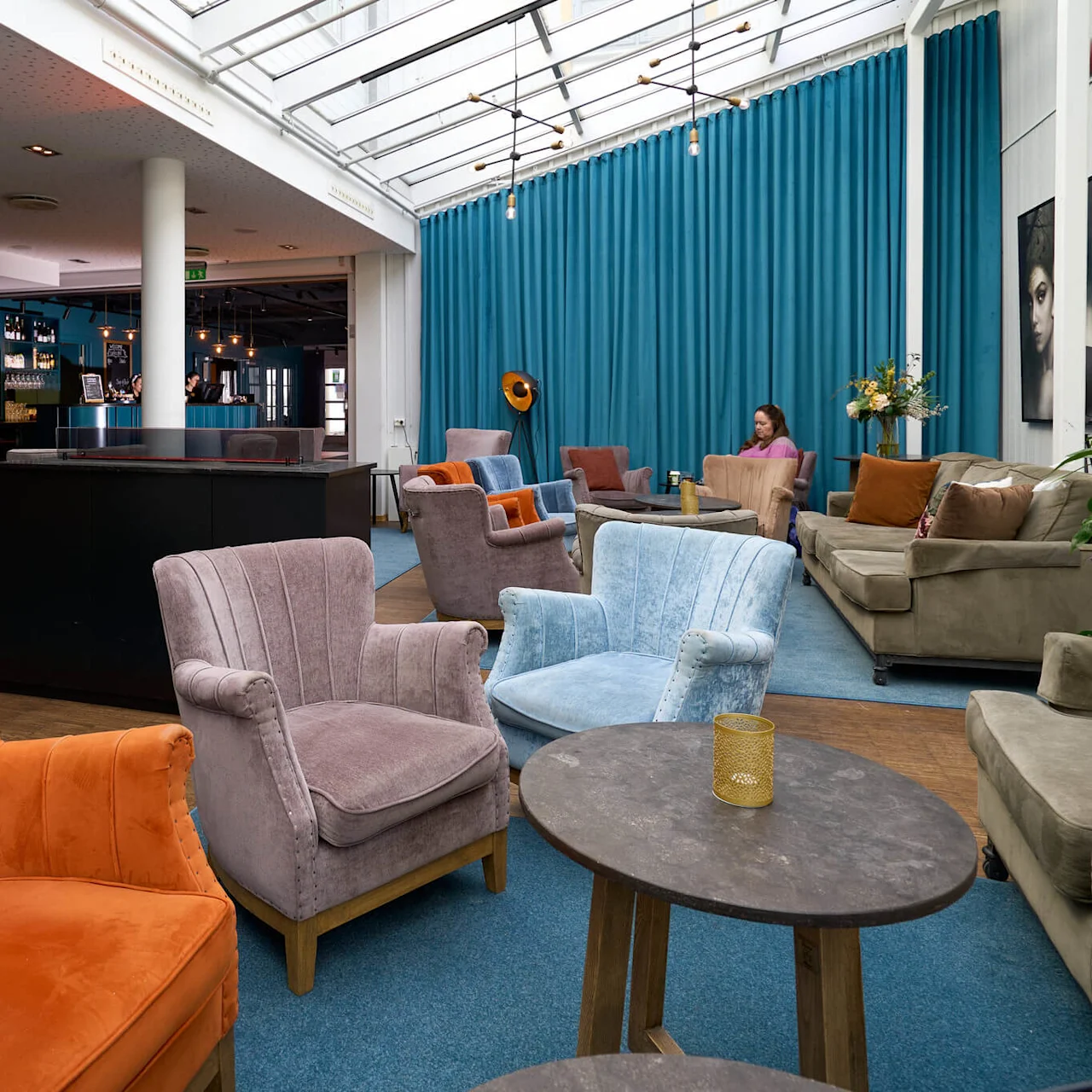 Farverige lænestole er placeret omkring borde i en veloplyst hotellobby med en bar og en person, der arbejder ved en bærbar computer i baggrunden.