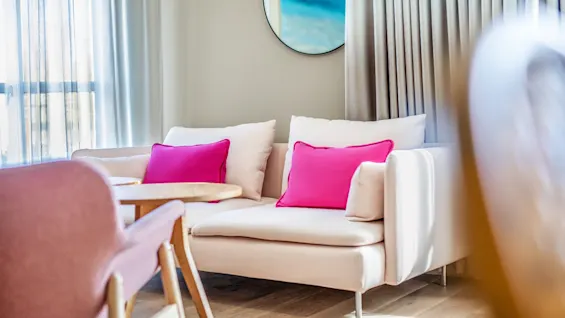 Cream sofa with pink cushions sits in a bright living room, adjacent to a wooden table and chairs. Curtains and a round mirror adorn the wall.