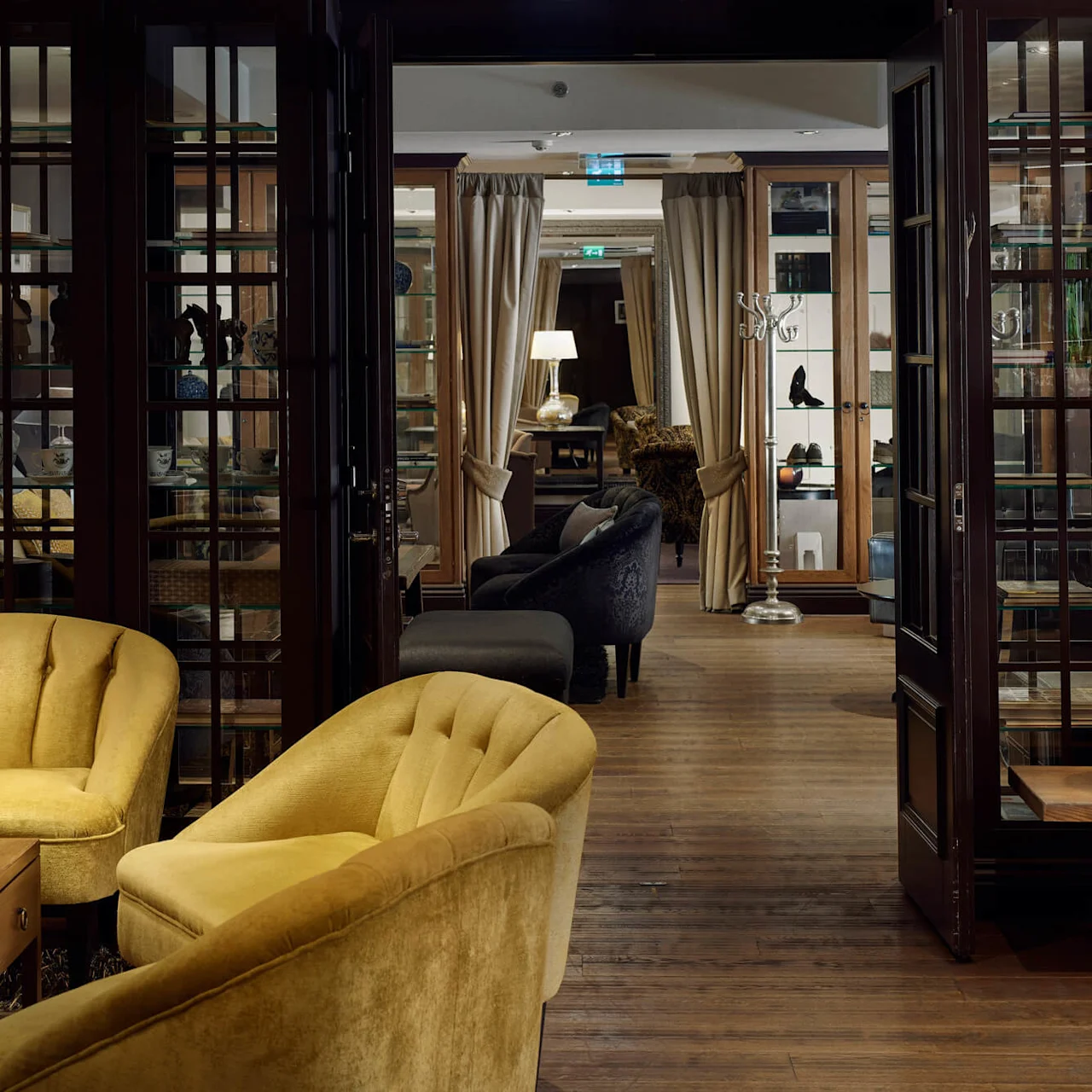 Yellow armchairs sit near a wooden coffee table with a newspaper and glass, in a warmly lit room with bookshelves and soft lighting.