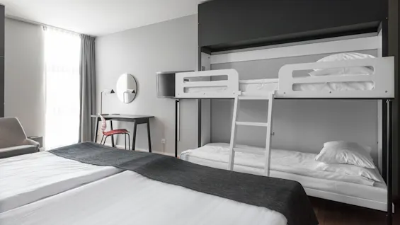 Bunk bed with white linens sits against a wall. A ladder connects the beds. A nearby work desk has a chair, lamp, and round mirror. A window illuminates the room.