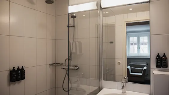 Ett väl upplyst badrum på Home Hotel Grand, Helsingborg, som visar en kaklad dusch och ett handfat med en spegel som reflekterar ett rum.