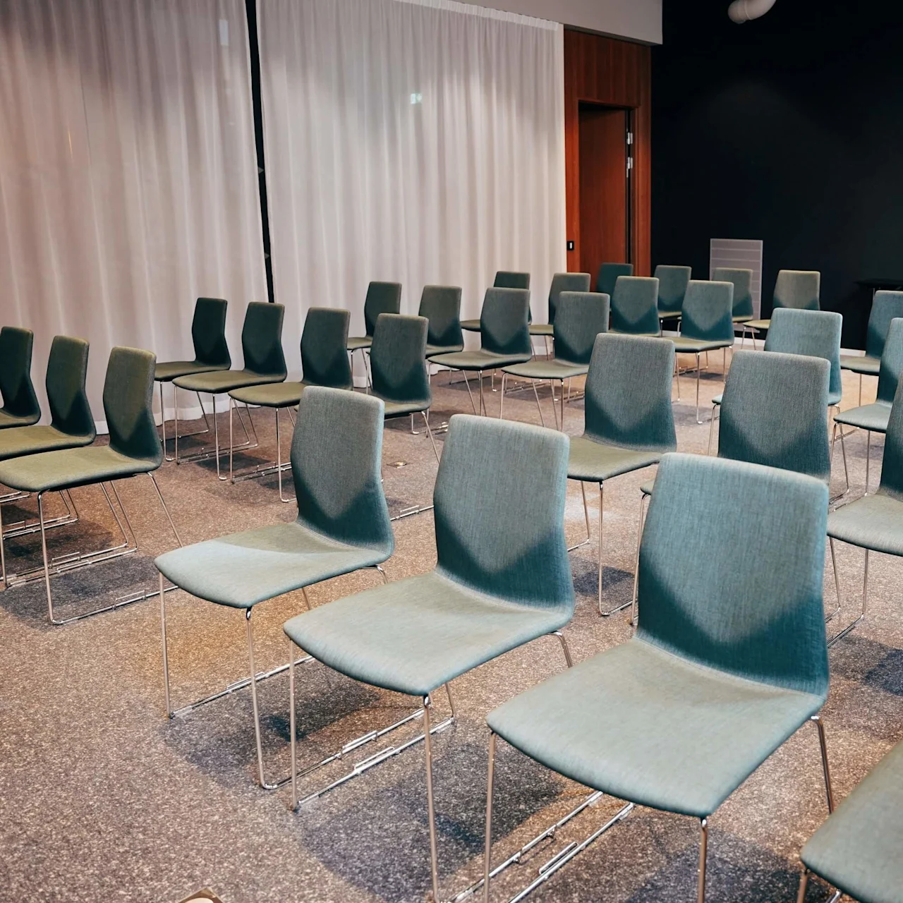 Rows of empty, gray chairs are arranged in a conference room with gray carpet and white curtains. A clock hangs on the dark wall.