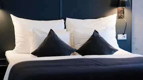 Bed with white and dark pillows is neatly arranged. Wall lamps illuminate the headboard, while surrounding concrete walls create a minimalist, modern atmosphere.