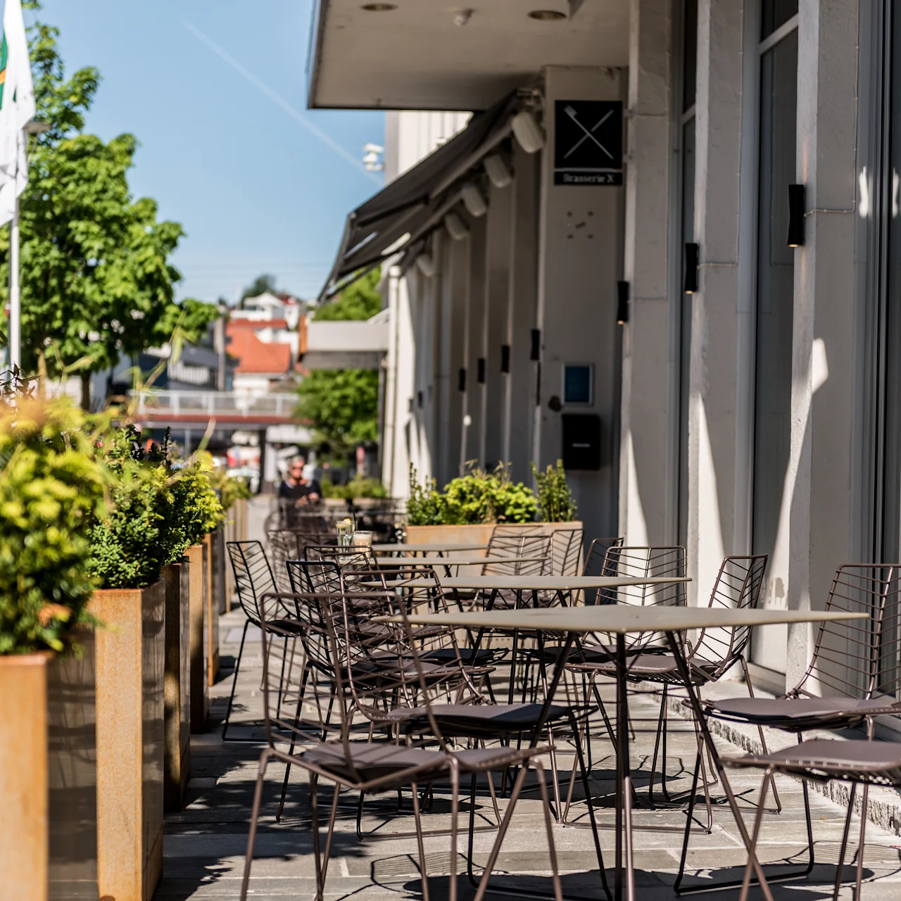 Ulkona kahvilapöydät ja tuolit reunustavat aurinkoista jalkakäytävää vehreiden istutusten vieressä kirkkaan sinisen taivaan alla. Kyltti lukee "Brasserie X."