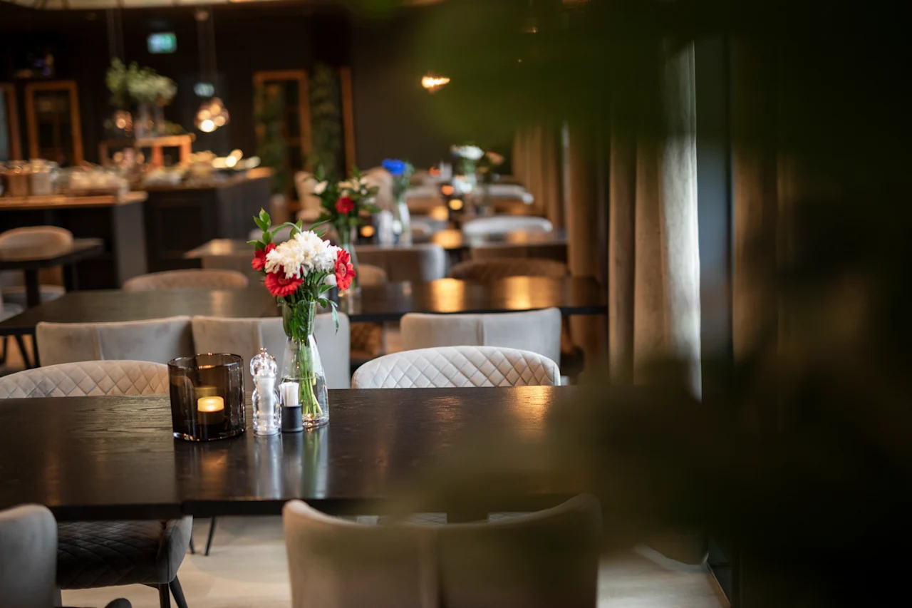 Et hyggeligt restaurantinteriør med borde dækket til spisning, med blomsterarrangementer og varm belysning.