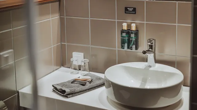 Handfat med kran placerad på en vit bänk. Två väggmonterade tvåldispensrar ovanför, bredvid vikta handdukar och glas. Beige kakel runtomkring. Text: "THINK SUSTAINABLY."