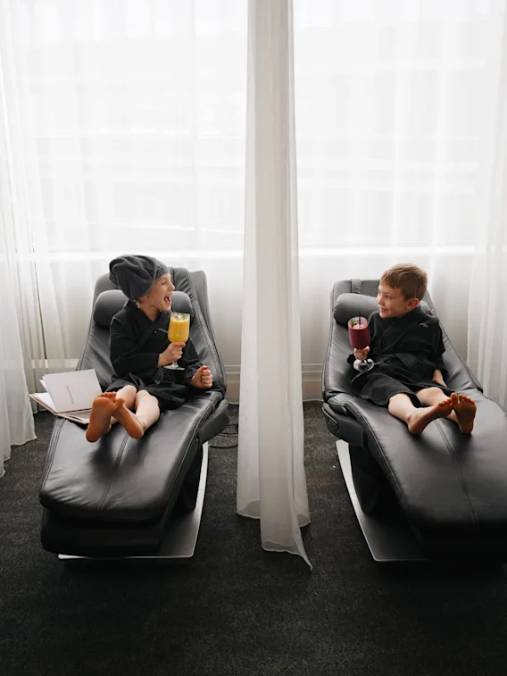 Two children in bathrobes relaxing on lounge chairs and enjoying colorful drinks at a spa